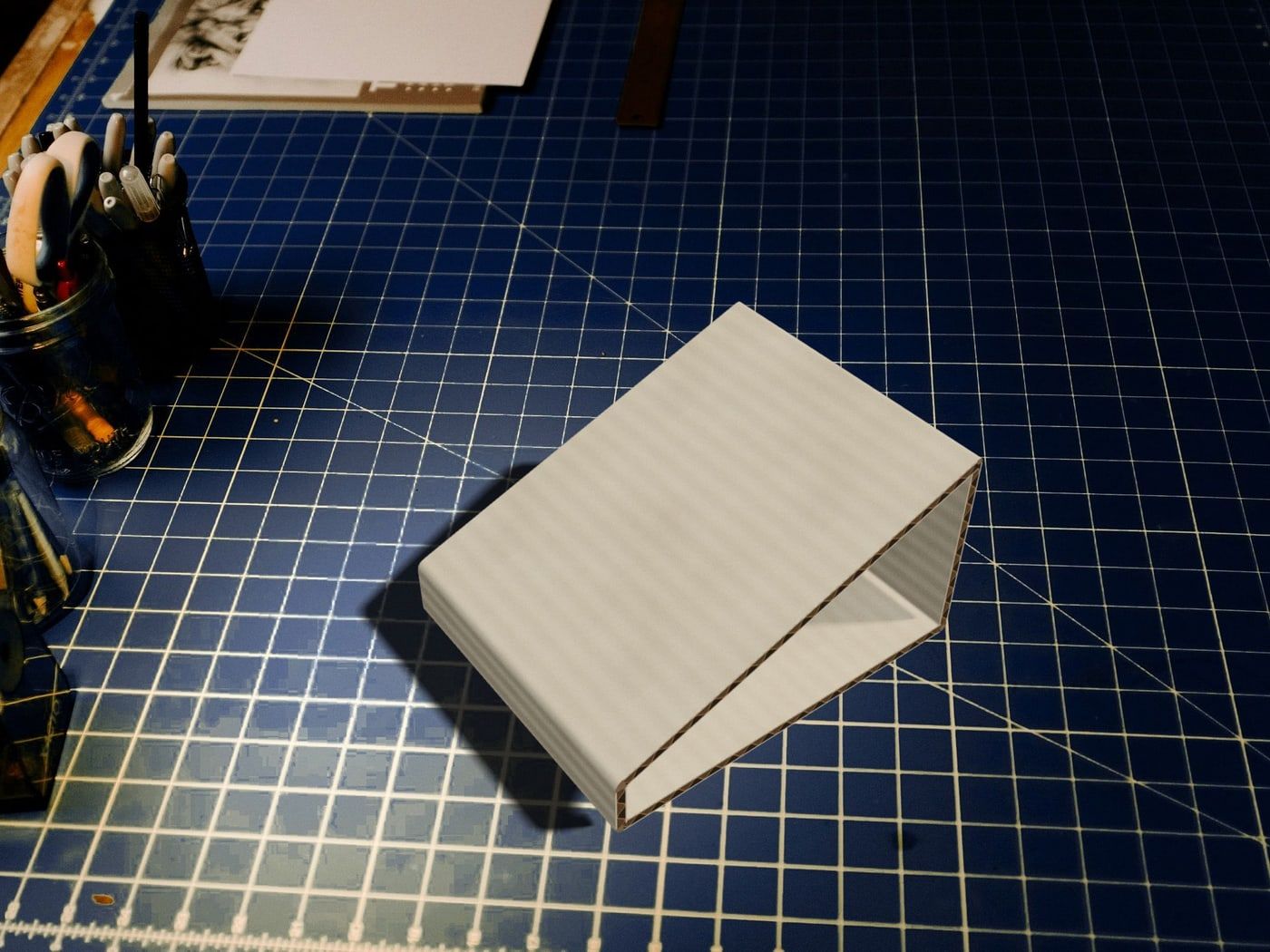 An off-white corrugated-cardboard prototype of the tip tent on a dark blue cutting mat in a design studio, beside jars of pencils and markers.